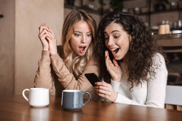 Эмоциональные шокированные девушки друзья сидят в кафе с помощью мобильного телефона . — стоковое фото