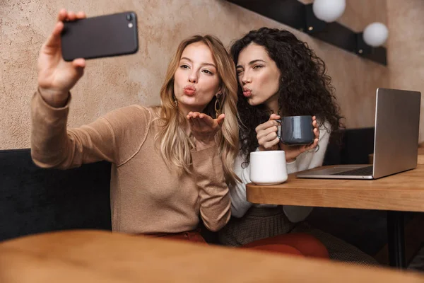 Dizüstü bilgisayar kullanarak kafede oturan heyecanlı mutlu kız arkadaşlar telefonla selfie çekmek. — Stok fotoğraf