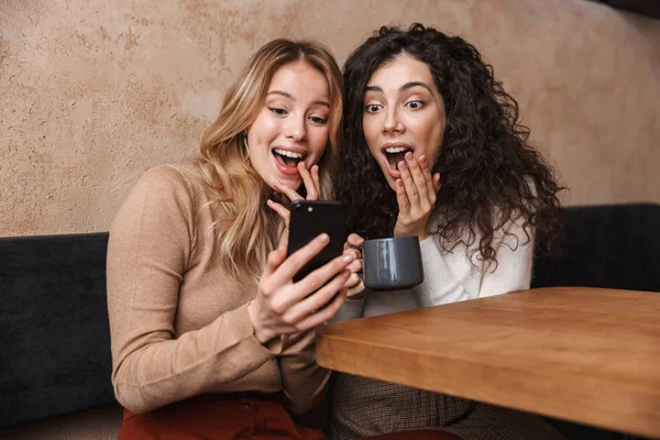 Эмоциональные шокированные девушки друзья сидят в кафе с помощью мобильного телефона . — стоковое фото
