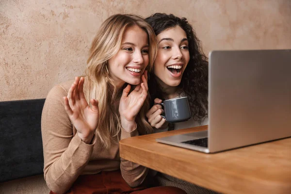 Gelukkig mooie meisjes vrienden zitten in cafe met behulp van laptop computer zwaaien praten. — Stockfoto