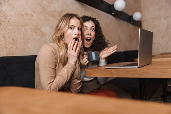 Эмоциональные шокированные девушки друзья сидят в кафе с помощью ноутбука . — стоковое фото