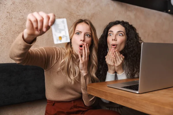 Sorprendido chicas guapas amigos sentados en la cafetería bebiendo café usando ordenador portátil con tarjeta de crédito . — Foto de Stock