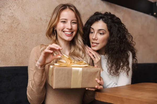 Ενθουσιασμένοι ευτυχισμένοι όμορφα κορίτσια φίλοι κάθονται στο καφέ κρατώντας παρόν κουτί δώρου. — Φωτογραφία Αρχείου