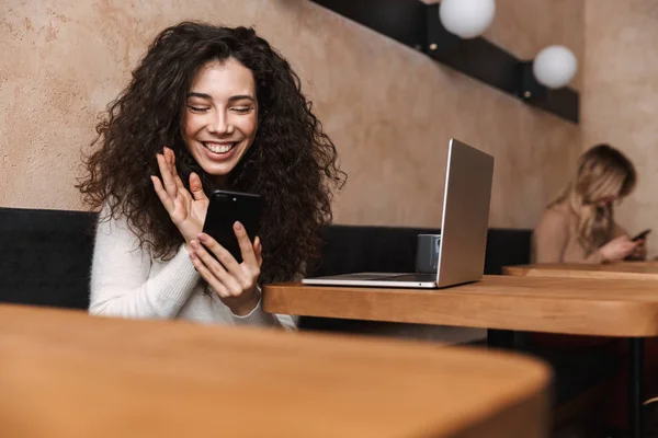 Mutlu kız dizüstü bilgisayar ve cep telefonu kullanarak kafede oturan. — Stok fotoğraf