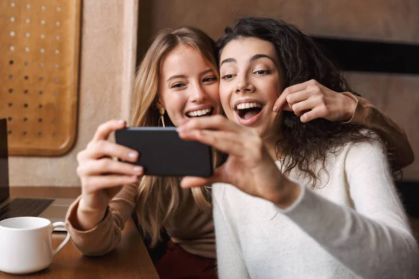 Heyecanlı mutlu güzel kız arkadaşlar cep telefonu kullanarak kafede oturan. — Stok fotoğraf