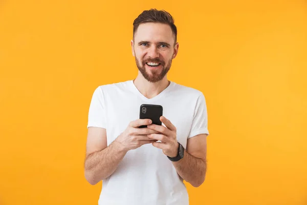 Gelukkig jonge knappe man poseren geïsoleerd over gele muur achtergrond met behulp van mobiele telefoon. — Stockfoto