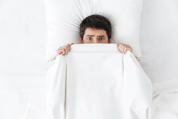 Scared handsome young man in the morning under blanket hide in bed.