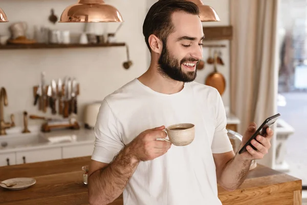 Portrait of laughing brunette man 20s drinking coffee and using — Stock Photo, Image