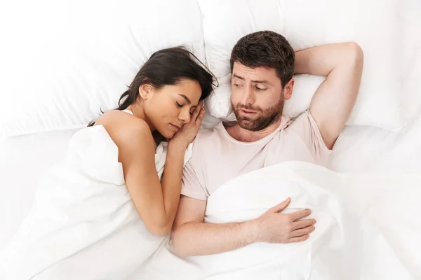 Descontento joven confundido se encuentra en la cama debajo de la manta cerca de la mujer dormida . —  Fotos de Stock