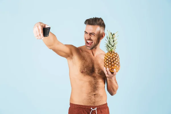 Knappe opgewonden gelukkige volwassen man in badmode poseren geïsoleerd over blauwe muur achtergrond Neem een selfie per telefoon met ananas. — Stockfoto