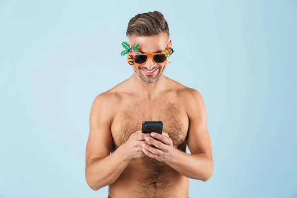 Emocionado hombre adulto feliz en traje de baño posando aislado sobre fondo de pared azul utilizando el teléfono móvil . — Foto de Stock