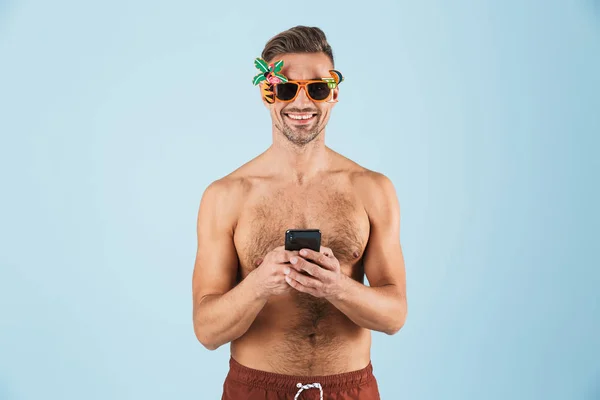 Возбужденный счастливый взрослый мужчина в купальниках позирует изолированно на фоне синей стены с помощью мобильного телефона . — стоковое фото
