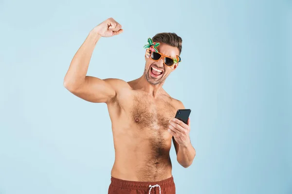 Emocionado hombre adulto feliz en traje de baño posando aislado sobre fondo de pared azul utilizando el teléfono móvil . — Foto de Stock