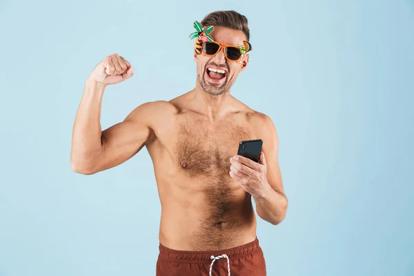 Ενθουσιασμένος ευτυχισμένος ενήλικος άνθρωπος σε μαγιό που παρουσιάζουν απομονωμένα πάνω από μπλε τοίχο φόντο χρησιμοποιώντας το κινητό τηλέφωνο. — Φωτογραφία Αρχείου