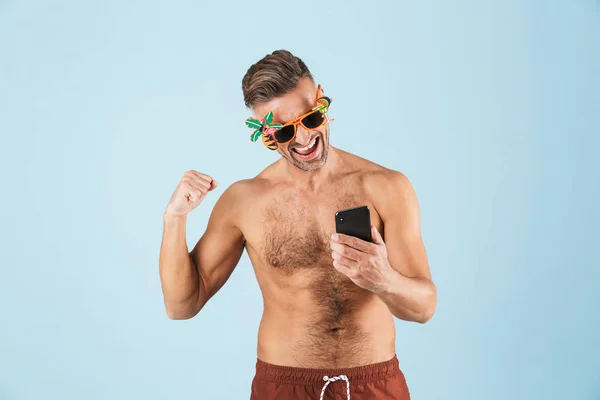 Emocionado hombre adulto feliz en traje de baño posando aislado sobre fondo de pared azul utilizando el teléfono móvil . — Foto de Stock