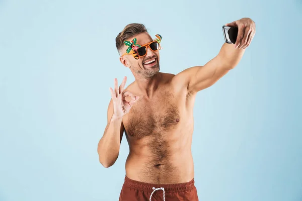 Emocionado hombre adulto feliz en traje de baño posando aislado sobre fondo de pared azul utilizando el teléfono móvil que muestra gesto bien tomar una selfie . —  Fotos de Stock