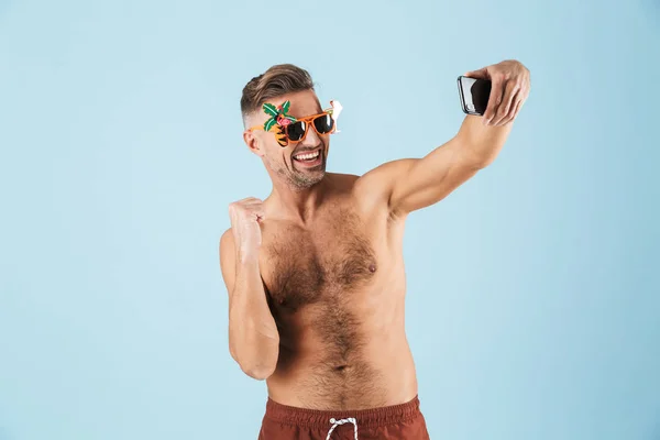 Emocionado hombre adulto feliz en traje de baño posando aislado sobre fondo de pared azul usando el teléfono móvil tomar una selfie . —  Fotos de Stock