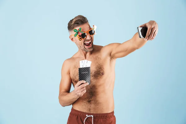 Emocionado hombre adulto feliz en traje de baño posando aislado sobre fondo de pared azul usando el teléfono móvil tomar una selfie . — Foto de Stock