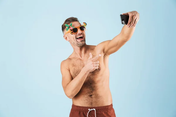 Emocionado hombre adulto feliz en traje de baño posando aislado sobre fondo de pared azul usando el teléfono móvil tomar una selfie . — Foto de Stock