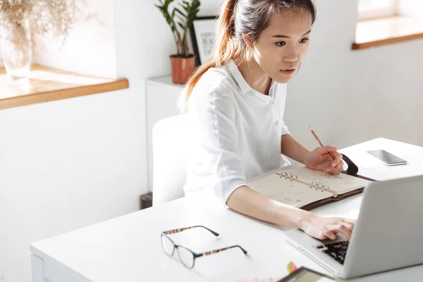 Geconcentreerde Aziatische zakenvrouw iets schrijven en met behulp van laptop computer — Stockfoto