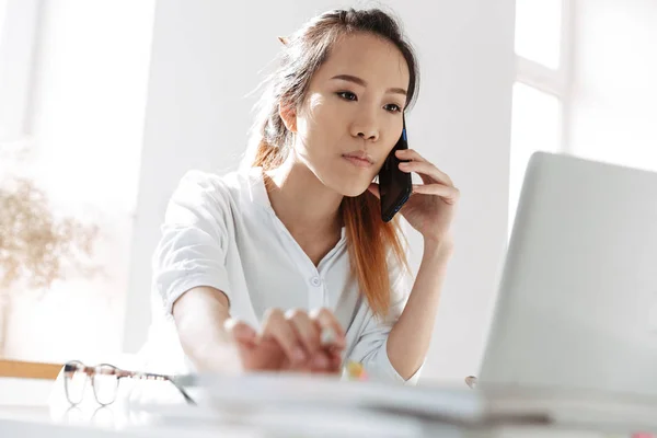 Serieuze Aziatische zakenvrouw praten door smartphone en het gebruik van laptop — Stockfoto