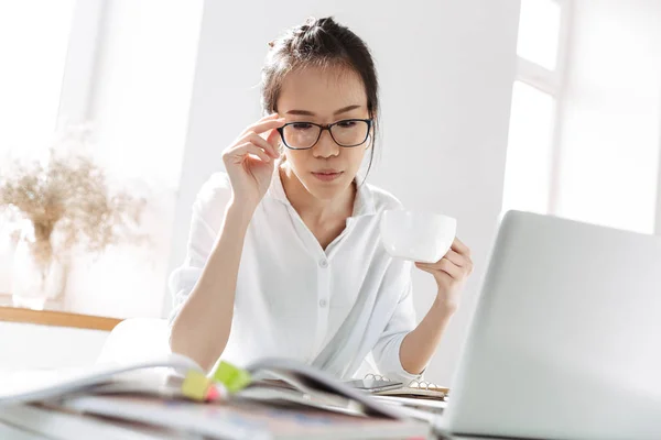 Serious asian business woman in eyeglasses drinking coffee