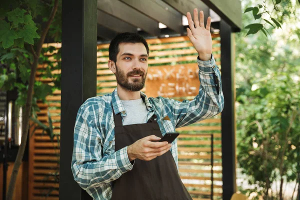 Foto de tipo amigable barista sonriendo mientras usa el teléfono inteligente en —  Fotos de Stock