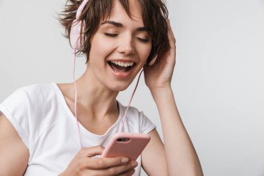 Genç, oldukça heyecanlı, mutlu bir kadın, beyaz duvar arkasında izole bir şekilde poz veriyor. Kulaklıklı, cep telefonlu müzik dinliyor..