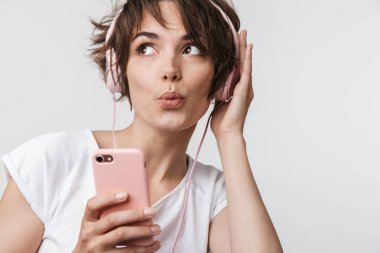Genç, oldukça heyecanlı, mutlu bir kadın, beyaz duvar arkasında izole bir şekilde poz veriyor. Kulaklıklı, cep telefonlu müzik dinliyor..