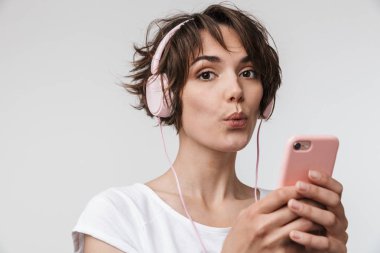 Genç, oldukça heyecanlı, mutlu bir kadın, beyaz duvar arkasında izole bir şekilde poz veriyor. Kulaklıklı, cep telefonlu müzik dinliyor..