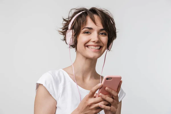 Jovem mulher feliz muito animado posando isolado sobre fundo da parede branca usando telefone celular ouvir música com fones de ouvido . — Fotografia de Stock