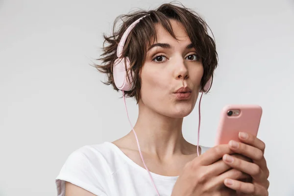 Молодая довольно взволнованная счастливая женщина позирует изолированы на белом фоне стены с помощью мобильного телефона прослушивания музыки с наушниками . — стоковое фото