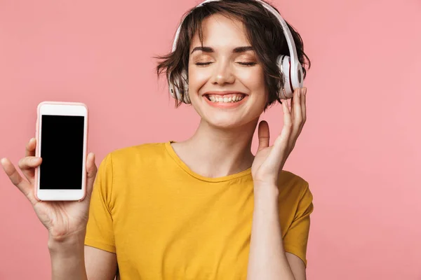 Vacker kvinna poserar isolerade över rosa vägg bakgrund lyssna musik med hörlurar med mobiltelefon som visar displayen. — Stockfoto