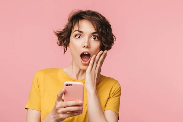 Joven mujer hermosa impactada posando aislada sobre fondo de pared rosa usando teléfono móvil . — Foto de Stock