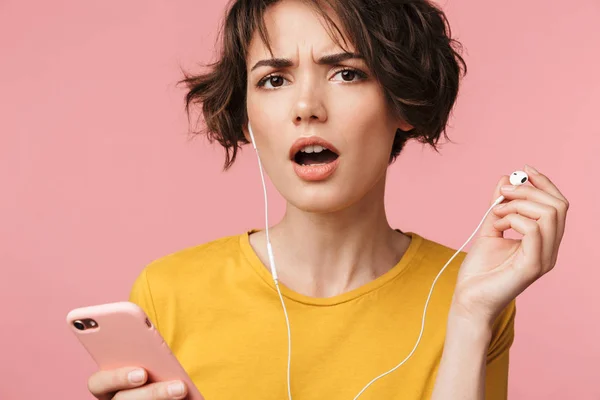 Mujer hermosa joven concentrada posando aislada sobre fondo de pared rosa escuchando música con auriculares usando el teléfono móvil tratar de oírte . —  Fotos de Stock
