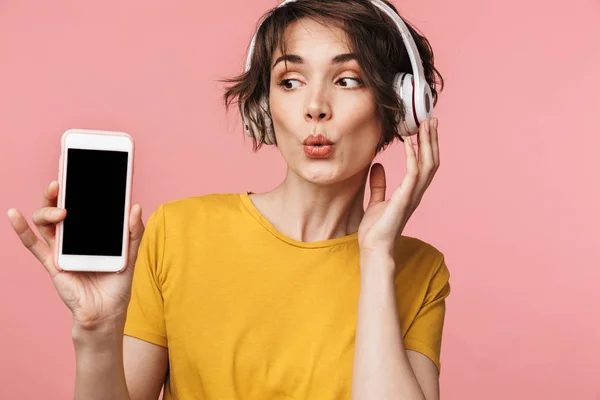 Bella donna posa isolata su sfondo rosa muro ascoltare musica con auricolari utilizzando il telefono cellulare che mostra display . — Foto Stock