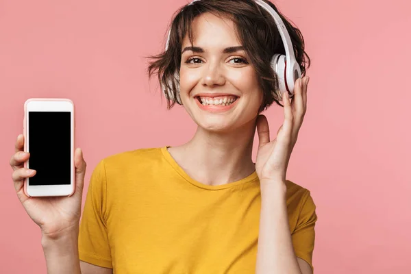 Bella donna posa isolata su sfondo rosa muro ascoltare musica con auricolari utilizzando il telefono cellulare che mostra display . — Foto Stock