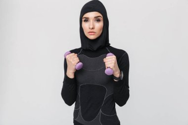 Genç kadın fitness müslümanı, beyaz duvar arkasına izole edilmiş halterlerle egzersiz yapıyor..