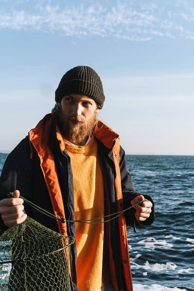 Bello concentrato giovane pescatore indossa cappotto e cappello in riva al mare . — Foto Stock