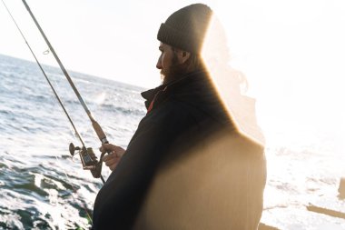Yakışıklı genç adam balıkçı sahilde ceket ve şapka giyiyor.