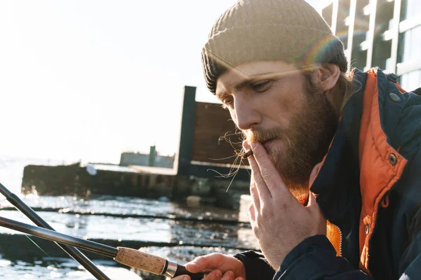 Bello giovane pescatore che indossa cappotto e cappello al mare fumare . — Foto Stock