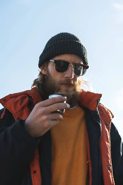 Schöner junger Mann Fischer mit Mantel und Hut am Strand, der heißen Tee trinkt. — Stockfoto