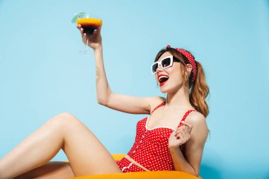 Mayo giymiş genç bir kız şişme yüzükle mavi arka planda yalnız poz veriyor ve kokteyl içiyor..