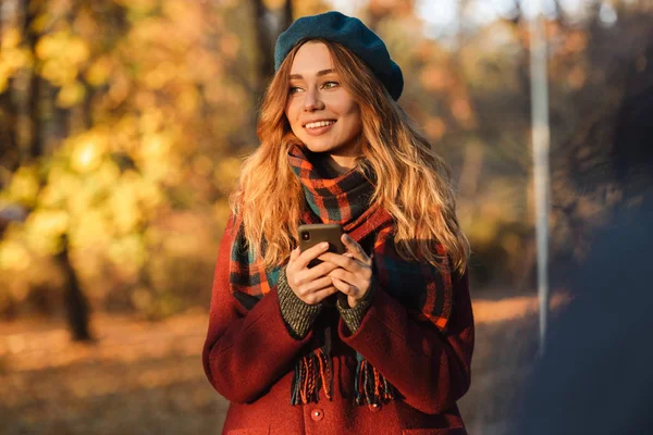 Happy young pretty woman walking outdoors in autumn spring park using mobile phone chatting. — Stock Photo, Image
