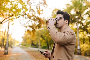 Şık esmer adam portresi akıllı telefon üzerinde ceket yazarak ve sonbahar parkta paket kahve içme
