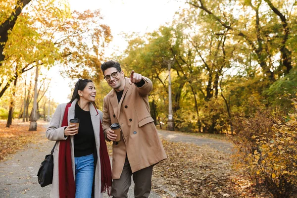 Retrato de pareja feliz tomando café para llevar de tazas de papel mientras camina en el parque de otoño —  Fotos de Stock