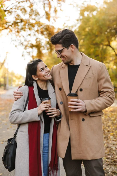 Retrato de pareja joven tomando café para llevar y mirándose mientras caminan en el parque de otoño —  Fotos de Stock