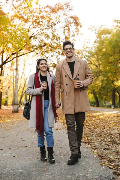 Retrato de pareja casual tomando café para llevar de tazas de papel mientras camina en el parque de otoño —  Fotos de Stock