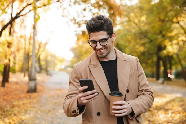 Imagen de hombre casual guapo usando abrigo usando teléfono celular y sonriendo n parque de otoño —  Fotos de Stock