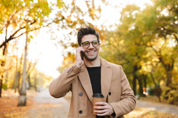Wizerunek przystojnego młodego mężczyzny noszący płaszcz rozmawiający na smartfonie i uśmiechnięty w jesiennym parku — Zdjęcie stockowe